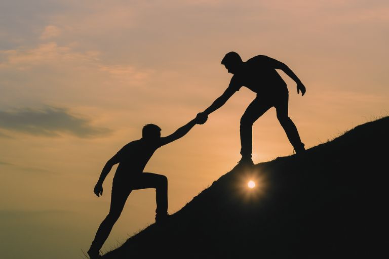 Silhouette of someone reaching out to another person to help them up a steep hill